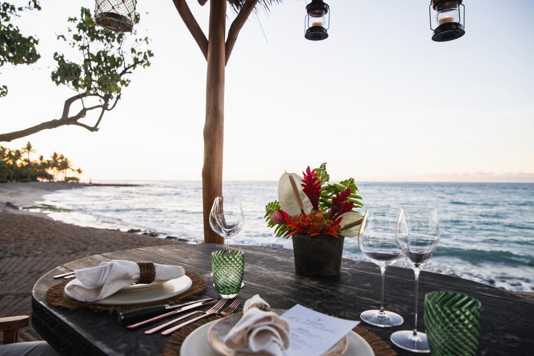 A dining table set for two with wine glasses, plates, and a flower arrangement on the beach.